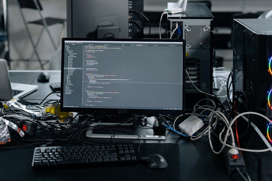 A person with light skin is working on a laptop placed on a dark, reflective tabletop. The laptop screen displays lines of code in a text editor with a dark background and colorful syntax highlighting